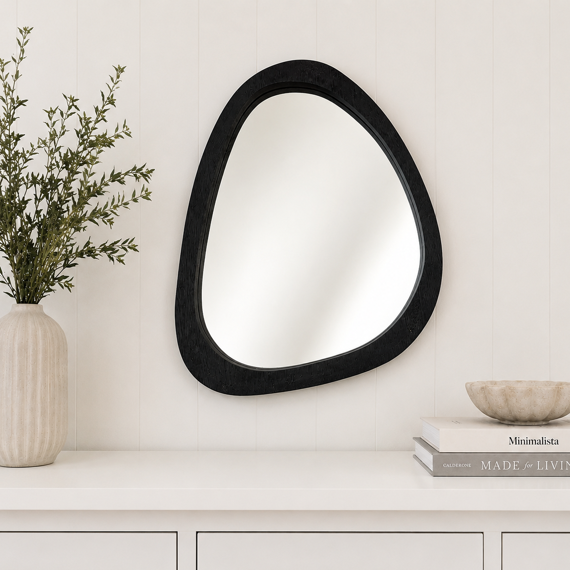 Black abstract mirror on a white wall with a vase and books below.