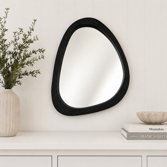 Black abstract mirror on a white wall with a vase and books below.